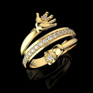 Könnte beinhalten: Ein goldfarbener Ring mit einem einzigartigen Design. Der Ring zeigt eine Hand, die einen Diamanten hält, ein Band aus Diamanten und einen Fuß mit einem Diamanten. Der Ring ist vor einem schwarzen Hintergrund platziert.