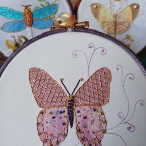 May include: A pink and gold butterfly with beaded accents is embroidered on a white fabric stretched in a wooden embroidery hoop.