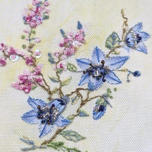 May include: Detailed embroidery of flowers in shades of light blue and pink, with green leaves and gold-coloured stems. The flowers are adorned with beads and sequins, set against a cream background.