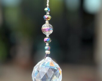 Op de afbeelding: Een hangend zonnevanger-ornament met een grote, heldere, gefacetteerde kristallen bol die aan een zilverkleurige metalen ketting hangt. De ketting is versierd met kleinere, iriserende, regenboogkleurige kralen en heldere glazen tussenstukjes.