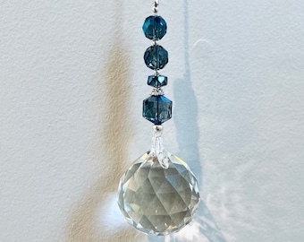 Op de afbeelding: Een kristallen zonenvanger met een heldere, gefacetteerde bal en een zilveren ketting. De ketting is versierd met blauwe kristallen kralen.