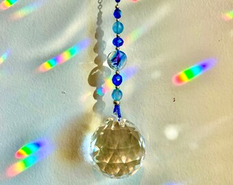 Puede incluir: Una bola de cristal colgando de una cadena plateada con cuentas azules y transparentes. La bola de cristal proyecta arcoíris en la pared blanca.