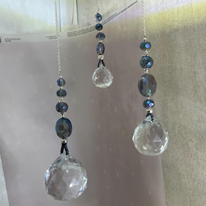 May include: Three hanging crystal suncatchers. Each features a faceted crystal ball at the bottom, with a series of blue and silver beads strung above. The crystals and beads are suspended by silver chains.