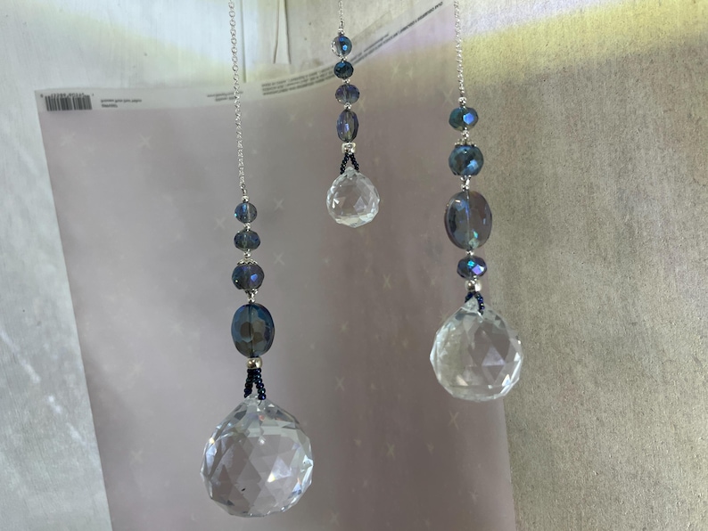 May include: Three hanging crystal suncatchers. Each features a faceted crystal ball at the bottom, with a series of blue and silver beads strung above. The crystals and beads are suspended by silver chains.