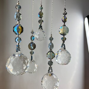 May include: Five hanging crystal suncatchers with faceted spheres and beads in shades of clear, blue, and iridescent colors. The crystals are suspended from silver chains, creating a decorative and elegant display.