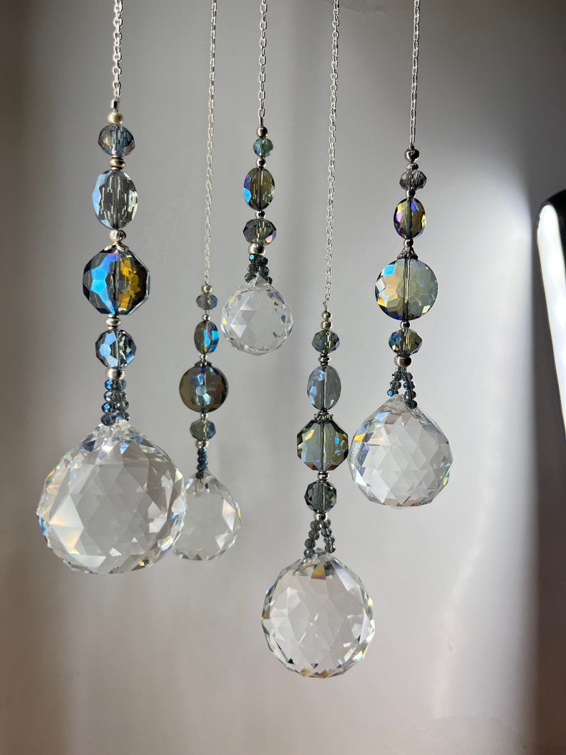 May include: Five hanging crystal suncatchers with faceted spheres and beads in shades of clear, blue, and iridescent colors. The crystals are suspended from silver chains, creating a decorative and elegant display.