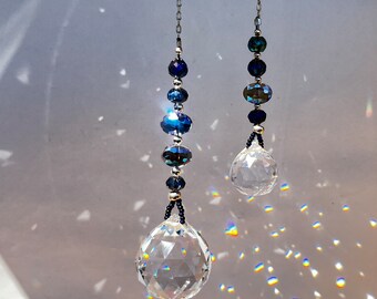 Puede incluir: Dos atrapasoles de cristal con cuentas azules colgando de cadenas plateadas. El atrapasol más grande tiene una bola de cristal más grande al final de la cadena. El atrapasol más pequeño tiene una bola de cristal más pequeña al final de la cadena.
