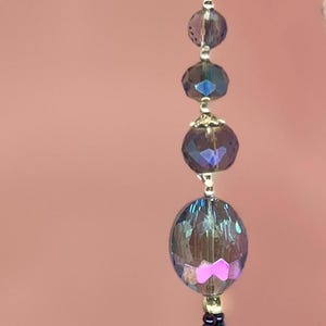 May include: A decorative crystal prism suncatcher with iridescent beads and a silver chain. The prism is faceted and hangs below a series of iridescent blue and purple beads. The background is a solid pink.