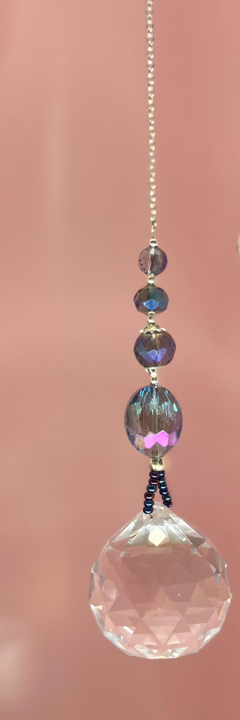 May include: A decorative crystal prism suncatcher with iridescent beads and a silver chain. The prism is faceted and hangs below a series of iridescent blue and purple beads. The background is a solid pink.