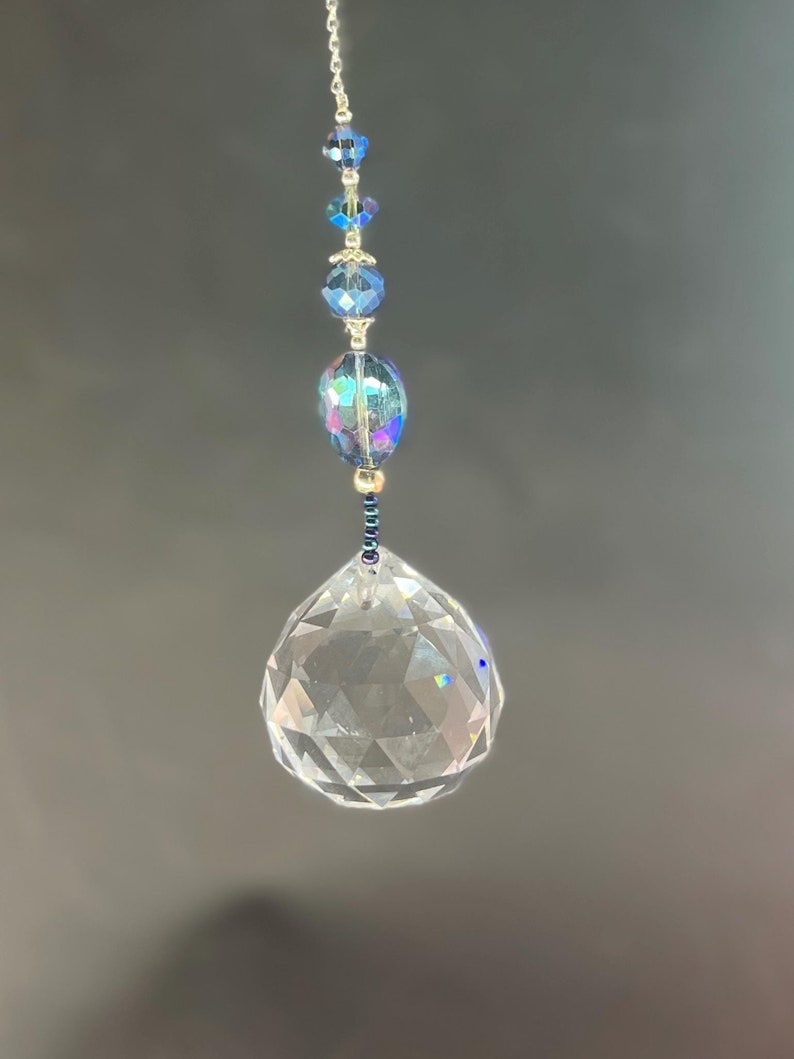 May include: A clear crystal ball suncatcher with a faceted surface, suspended by a silver chain. Above the ball are iridescent blue and clear crystal beads. The suncatcher is designed to catch and reflect light.