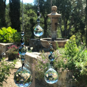 May include: Three hanging crystal suncatchers with faceted spheres and smaller beads. The suncatchers are suspended by dark metal chains, reflecting light and creating a rainbow effect. The background shows a blurred outdoor scene with greenery and a fountain.