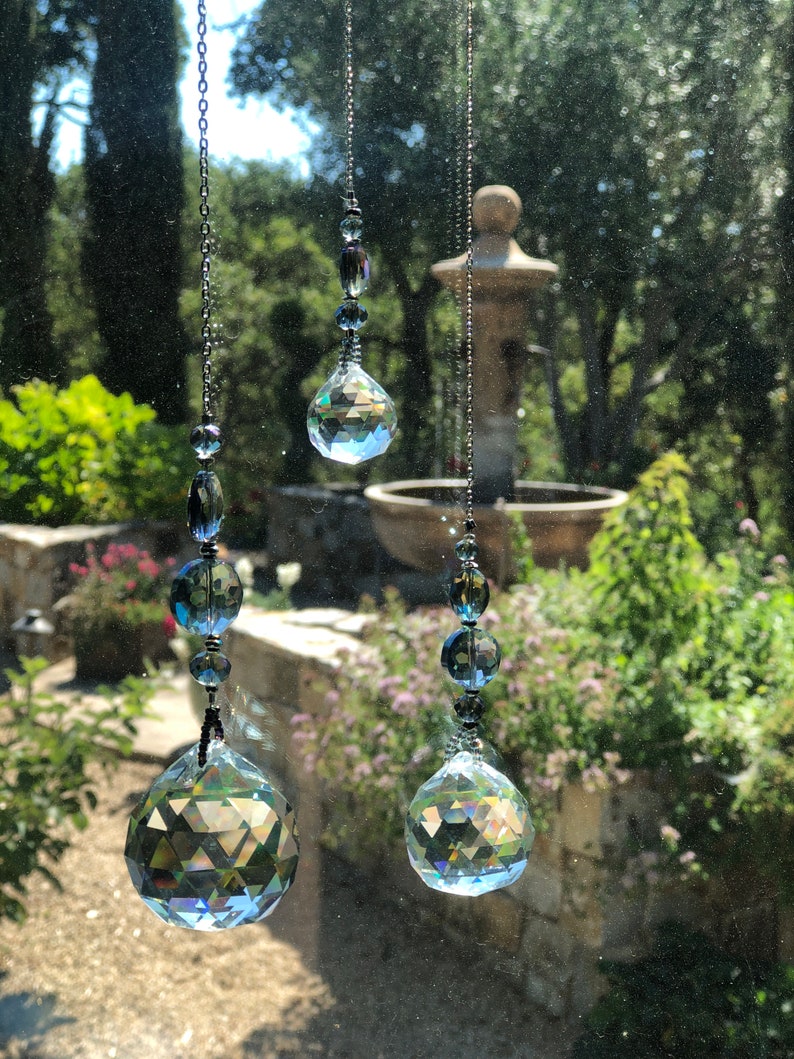May include: Three hanging crystal suncatchers with faceted spheres and smaller beads. The suncatchers are suspended by dark metal chains, reflecting light and creating a rainbow effect. The background shows a blurred outdoor scene with greenery and a fountain.