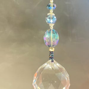 May include: A decorative crystal suncatcher with a faceted, clear crystal ball at the bottom. Above the ball are iridescent beads and smaller crystal spheres, strung on a gold-colored chain. The suncatcher hangs against a dark background.