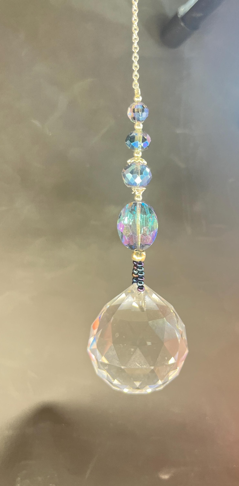 May include: A decorative crystal suncatcher with a faceted, clear crystal ball at the bottom. Above the ball are iridescent beads and smaller crystal spheres, strung on a gold-colored chain. The suncatcher hangs against a dark background.