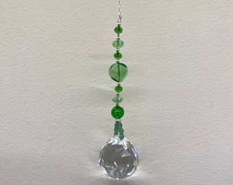 Op de afbeelding: Een kristallen zonenvanger met een heldere kristallen bol en groene glazen kralen die aan een zilveren ketting hangen.