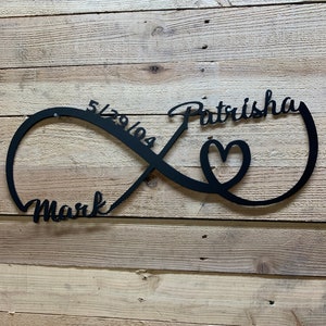 Puede incluir: Símbolo de infinito de metal negro con un corazón en el centro. Los nombres "Mark" y "Patricia" están a cada lado del símbolo, junto con la fecha "5.29.2021".