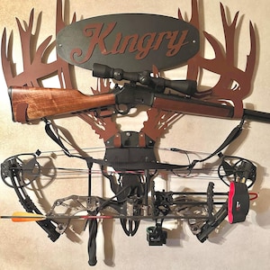 May include: A metal deer antler wall mount with a rifle and a compound bow hanging from it. The mount has a sign that says "Kingry".