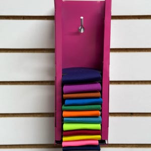 Puede incluir: Un estante de metal rosa con una pila de recortes de tela de colores. Los recortes de tela son de varios colores, incluyendo morado, naranja, azul, verde, amarillo y rosa.
