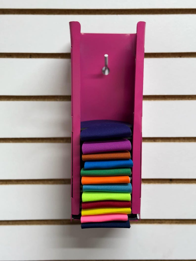 Puede incluir: Un estante de metal rosa con una pila de recortes de tela de colores. Los recortes de tela son de varios colores, incluyendo morado, naranja, azul, verde, amarillo y rosa.