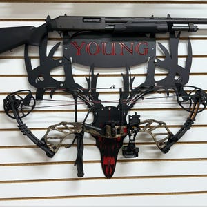 May include: A black shotgun and a camouflage compound bow are mounted on a black metal deer antler rack with the word "YOUNG" in red lettering.