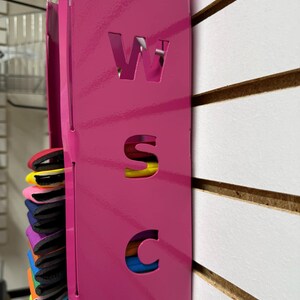 Puede incluir: Un organizador de pared de metal rosa con las letras W, S y C recortadas. El organizador tiene una ranura para guardar objetos, y hay objetos coloridos en la ranura.