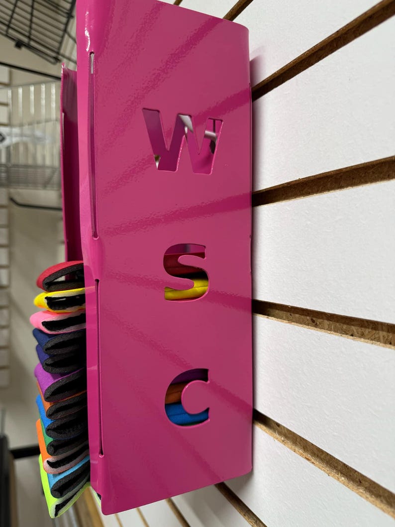 Puede incluir: Un organizador de pared de metal rosa con las letras W, S y C recortadas. El organizador tiene una ranura para guardar objetos, y hay objetos coloridos en la ranura.