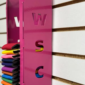 Puede incluir: Un organizador de pared de metal rosa con las letras "WSC" recortadas. El organizador tiene una ranura para guardar varias bandas de colores.