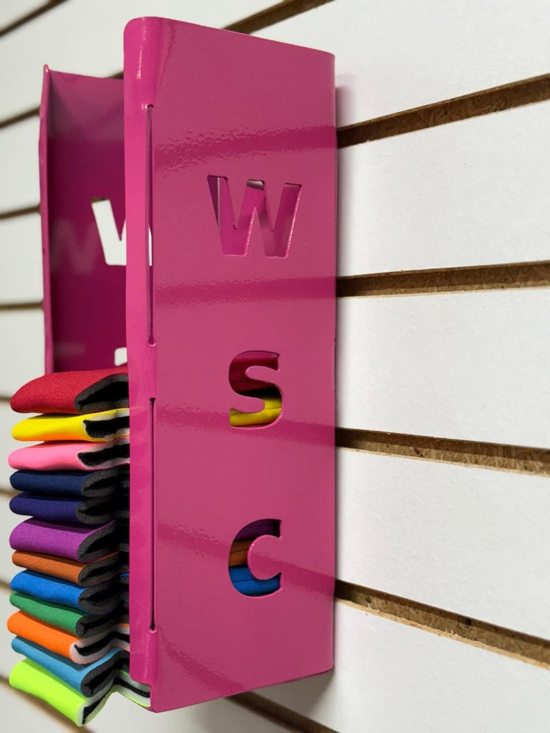 Puede incluir: Un organizador de pared de metal rosa con las letras "WSC" recortadas. El organizador tiene una ranura para guardar varias bandas de colores.