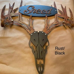 May include: Metal deer skull wall decor with antlers. The skull is black and the antlers are a rust color. The name "Snead" is written on a black oval plaque above the skull.