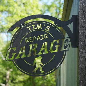 May include: Black metal sign for Tim's Repair Garage, featuring a circular design with the text "Tim's Repair Garage" and a silhouette of Bigfoot. The sign is mounted on a wooden post.