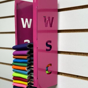 Puede incluir: Un organizador de pared de metal rosa con las letras "WSC" recortadas. El organizador tiene una ranura para guardar objetos y est&aacute; lleno de una pila de tiras de tela de colores.