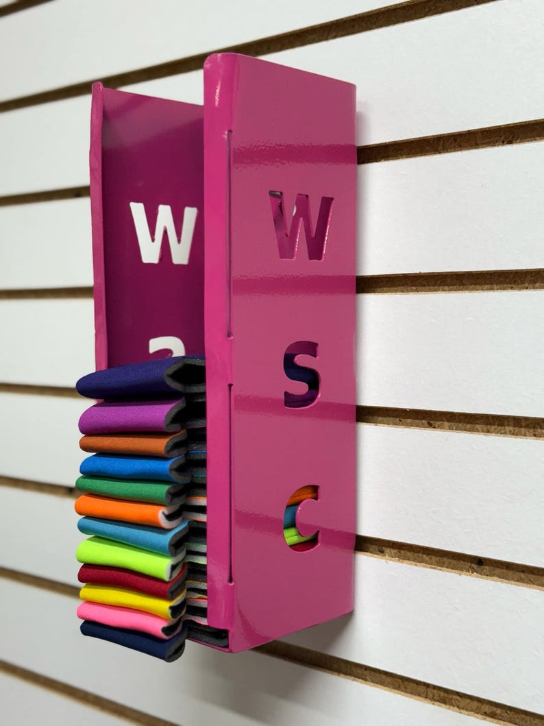 Puede incluir: Un organizador de pared de metal rosa con las letras "WSC" recortadas. El organizador tiene una ranura para guardar objetos y est&aacute; lleno de una pila de tiras de tela de colores.