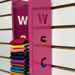 Puede incluir: Un organizador de pared de metal rosa con las letras "WSC" recortadas. El organizador tiene una ranura para guardar muestras de tela, que se muestran en una variedad de colores.