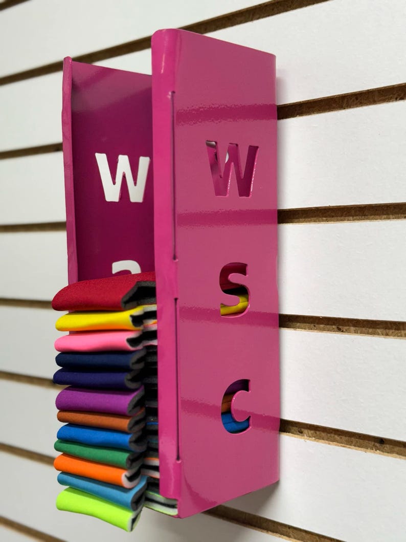 Puede incluir: Un organizador de pared de metal rosa con las letras "WSC" recortadas. El organizador tiene una ranura para guardar muestras de tela, que se muestran en una variedad de colores.
