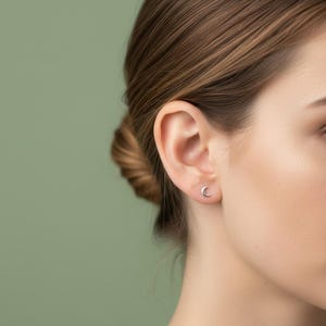 Puede incluir: Primer plano de una oreja con un pequeño pendiente plateado en forma de media luna. El pendiente tiene un diseño sencillo y minimalista. El fondo es verde liso y apagado.