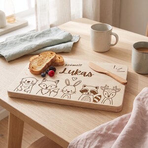Peut inclure: Une planche &agrave; d&eacute;couper rectangulaire en bois clair avec une gravure laser repr&eacute;sentant une loutre, un renard, un lapin, un raton laveur et un cerf. Elle porte l'inscription "kr&uuml;melt" et le pr&eacute;nom "Lukas" avec un petit c&oelig;ur, et un couteau en bois.