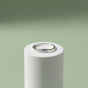 May include: A silver ring with the name "Logan" engraved on the front. The ring is displayed on a white cylindrical pedestal against a soft green background. The ring is a simple band design.