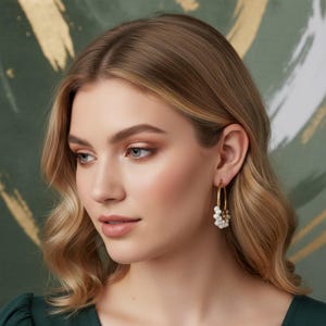 May include: A woman wearing gold hoop earrings with clusters of white pearls. The earrings are a classic design, adding a touch of elegance. The woman has blonde hair and is wearing a dark green top.