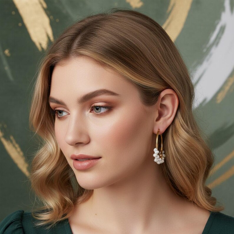 May include: A woman wearing gold hoop earrings with clusters of white pearls. The earrings are a classic design, adding a touch of elegance. The woman has blonde hair and is wearing a dark green top.