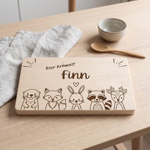 Peut inclure: Une planche &agrave; d&eacute;couper rectangulaire en bois clair avec une gravure laser repr&eacute;sentant cinq animaux de la for&ecirc;t : une loutre, un renard, un lapin, un raton laveur et un cerf. Le texte "Hier kr&uuml;melt Finn" est grav&eacute; au-dessus des illustrations.