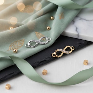 Könnte beinhalten: Zwei Unendlichkeits-Charm-Verbinder, einer silberfarben und einer goldfarben, jeweils mit klaren Kristallen besetzt. Die Charms werden auf einem salbeigrünen Stoff mit goldenen Blättern, einer schwarzen Schieferplatte und einer weißen Oberfläche mit einem grünen Band präsentiert.
