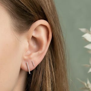 May include: A close-up of an ear with two silver bar earrings. The earrings are simple, minimalist designs, one longer than the other. The background is a soft green color, and the person's hair is brown.