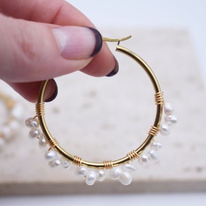May include: Gold-tone hoop earrings adorned with small, white freshwater pearls. The hoops are accented with gold wire wrapping. The earrings are held up against a soft, neutral background.