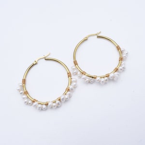 May include: A pair of gold-colored hoop earrings. Each earring features a gold hoop with a section wrapped in gold wire and adorned with small, white, teardrop-shaped pearls. The earrings are displayed against a white background.