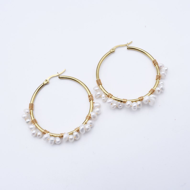 May include: A pair of gold-colored hoop earrings. Each earring features a gold hoop with a section wrapped in gold wire and adorned with small, white, teardrop-shaped pearls. The earrings are displayed against a white background.