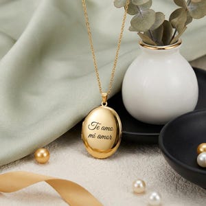 May include: A gold-coloured oval locket necklace inscribed with "Te amo mi amor". The pendant is suspended from a fine chain. The background features a white vase with greenery, gold-coloured pearls, and a black bowl.