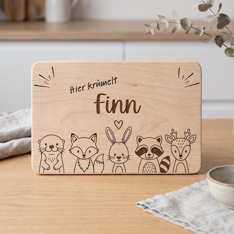 Peut inclure: Une planche &agrave; d&eacute;couper rectangulaire en bois clair avec un motif grav&eacute; au laser repr&eacute;sentant cinq animaux de la for&ecirc;t : une loutre, un renard, un lapin, un raton laveur et un cerf. Le texte "Hier kr&uuml;melt Finn" est grav&eacute; avec un petit c&oelig;ur.
