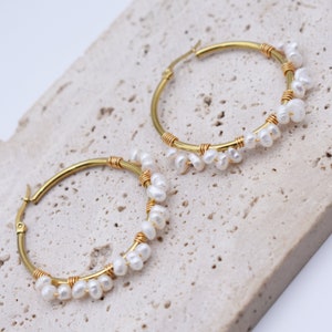 May include: A pair of gold-colored hoop earrings adorned with small, white pearls. The pearls are clustered along the bottom half of the hoops, secured with gold-colored wire. The earrings are displayed on a light-colored, textured surface.