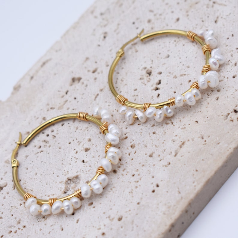 May include: A pair of gold-colored hoop earrings adorned with small, white pearls. The pearls are clustered along the bottom half of the hoops, secured with gold-colored wire. The earrings are displayed on a light-colored, textured surface.