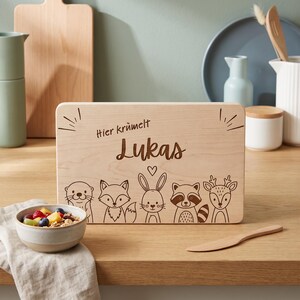 Peut inclure: Une planche &agrave; d&eacute;couper rectangulaire en bois clair avec une gravure laser repr&eacute;sentant cinq animaux des bois : une loutre, un renard, un lapin, un raton laveur et un cerf. Le texte "Hier kr&uuml;melt Lukas" est grav&eacute; au-dessus des illustrations.